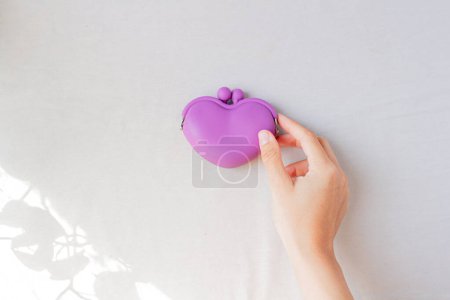 Photo for Female hand holding purple heart shaped coin wallet on white background, with some leaves shadow. - Royalty Free Image