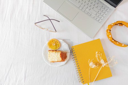 Foto de Inicio escritorio de oficina con ordenador portátil, pedazo de pastel naranja, planificador, vasos y flores secas sobre fondo blanco. - Imagen libre de derechos
