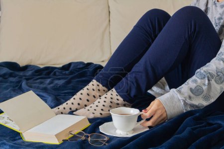 Téléchargez les photos : Jeune femme en pyjama, boire du thé et lire au lit - en image libre de droit