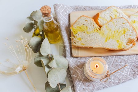 Foto de Vista superior del plato mediterráneo con aceite de oliva y pan. Una vela ligera, un eucalipto hojas secas, una tabla de cortar y un paño hacen un ambiente acogedor. - Imagen libre de derechos