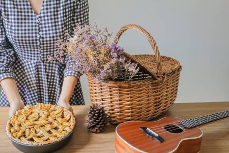 Téléchargez les photos : Composition d'automne avec des fleurs séchées dans le panier de pique-nique, ukulélé, tarte aux pommes décorée et jeune femme mettant tarte aux pommes sur la table - en image libre de droit