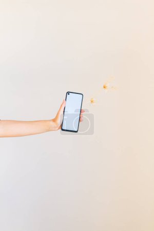 Téléchargez les photos : Jeune main féminine tenant un téléphone portable à écran vierge et quelques fleurs et feuilles séchées - en image libre de droit