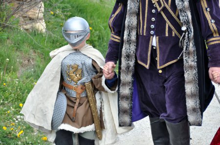 Photo for A young boy dressed as a knight, with a helmet on his face, a sword on his belt and a shield in his hand, is walking hand in hand with an adult wearing a robe. Spis Castle, Spissky Hrad, Slovakia. - Royalty Free Image