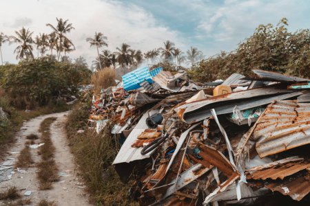 Foto de Un tiro de gran angular de un basurero ilegal en la selva; un montón de chatarra con pedazos de basura oxidados junto a un camino de tierra en entornos tropicales y palmeras en el fondo, isla Thoddoo, Maldivas - Imagen libre de derechos