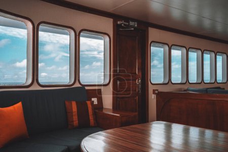 Photo for A row of windows with an exit door leading to a deck of a safari yacht; an interior of a cockpit with a seascape and cloudscape outside, and space of the wardroom with a table and sofa with cushions - Royalty Free Image