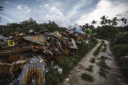 Foto de Foto de gran angular de un vertedero de basura ilegal ubicado en la selva cerca de un camino de tierra; un vertedero en un entorno tropical con muchas hojas de metal oxidado, bicicletas viejas y otra basura - Imagen libre de derechos