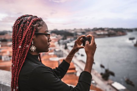 Téléchargez les photos : Une photo de profil d'une jeune voyageuse africaine avec des tresses rouges tirant sur une attraction touristique avec son smartphone ; une touriste noire photographiant le panorama du paysage urbain via son téléphone portable - en image libre de droit