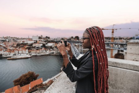 Téléchargez les photos : Vue latérale d'une charmante jeune femme africaine en tenue noire, avec de longues tresses rouges, filmant une image panoramique via son smartphone d'un paysage urbain avec une rivière et un coucher de soleil violet - en image libre de droit