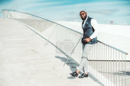Téléchargez les photos : Un entrepreneur afro-américain à barbe chauve mature à la mode dans un élégant costume formel sur mesure avec un gilet se penche sur la clôture dans la rue par un jour lumineux ; un espace de copie à gauche - en image libre de droit