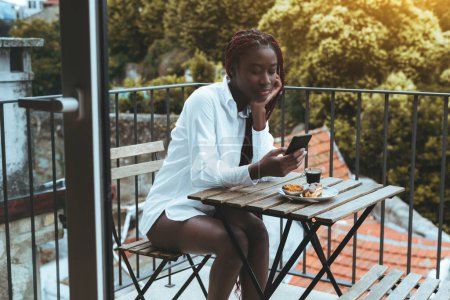 Foto de Una joven somnolienta negra con trenzas de caja roja está desplazando perezosamente una fuente de redes sociales en su teléfono inteligente mientras desayuna con un café expreso y pasteles en la terraza de su casa - Imagen libre de derechos