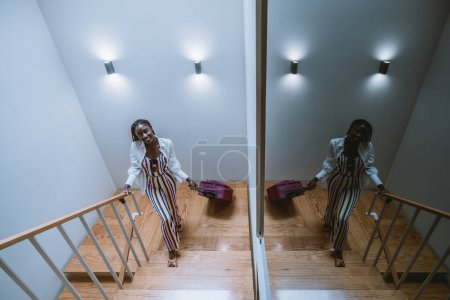 Foto de Una foto de gran angular de una joven encantadora viajera negra subiendo las escaleras con sus maletas de ruedas rosadas; una alegre joven africana está llegando a un alojamiento en un hotel en su viaje - Imagen libre de derechos