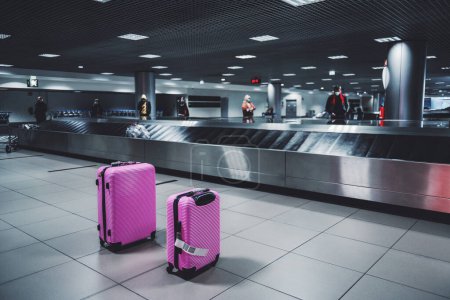 Téléchargez les photos : Focus sélectif sur deux valises à roulettes rose voyage bagages à côté de la courroie transporteuse de bagages dans une zone d'arrivée d'un terminal de l'aéroport et les gens qui attendent leurs bagages dans un arrière-plan déconcentré - en image libre de droit