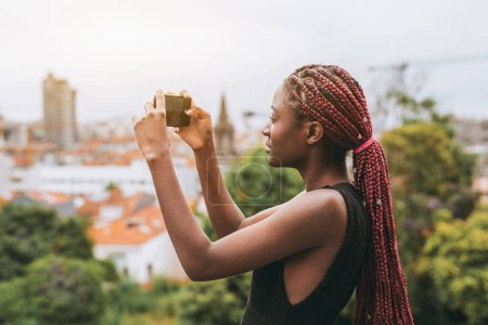Téléchargez les photos : Une photo de profil d'une jeune touriste noire prenant une photo panoramique du paysage urbain via son téléphone depuis le haut ; une jeune femme africaine filmant un panorama avec son téléphone portable - en image libre de droit