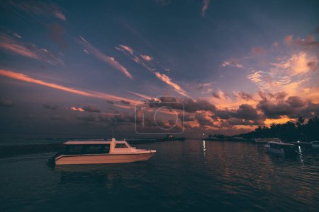 Foto de Vista panorámica de un embarcadero junto a una isla tropical con una espectacular puesta de sol en el fondo; un muelle oceánico con múltiples lanchas rápidas y barcos de transbordador con un impresionante paisaje nublado nocturno - Imagen libre de derechos