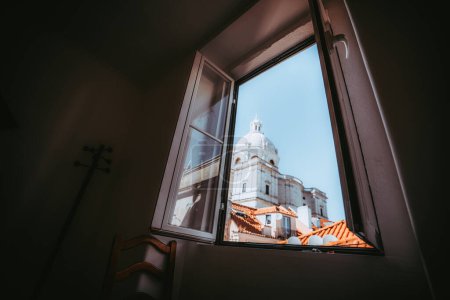 Foto de Una vista de gran angular desde la habitación doméstica oscura a través de la ventana abierta de una catedral con una cúpula, techos de azulejos de color naranja de las casas de Lisboa, y un cielo azul claro; enfoque selectivo en las persianas - Imagen libre de derechos
