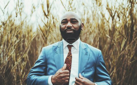 Téléchargez les photos : Portrait d'un homme noir mature chauve à la mode entrepreneur avec une barbe épaisse et dans un élégant costume bleu avec une cravate ajuste le blazer tout en se tenant à l'extérieur avec des roseaux en arrière-plan - en image libre de droit