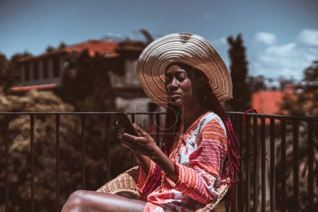 Foto de Retrato de una joven y elegante hembra negra con un vestido de verano y un sombrero de ala ancha, usando un teléfono inteligente mientras está sentada en un balcón; una hembra africana en un vestido desnudo llamando al aire libre - Imagen libre de derechos