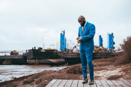 Photo for A fashionable bald mature black man is looking at his wristwatch, checking the time, while standing on a wooden pier with three metal supports in the background of the same blue color as his suit - Royalty Free Image