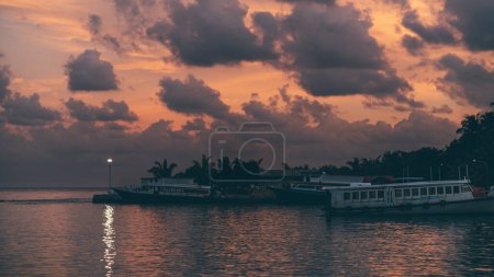 Téléchargez les photos : Un magnifique coucher de soleil lilas avec un paysage nuageux spectaculaire se reflétant dans l'eau de l'océan ondulé à côté de la jetée avec plusieurs ferry-boats et autres navires amarrés près de l'île tropicale Thoddoo, Maldives - en image libre de droit