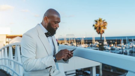 Téléchargez les photos : Une vue de profil d'un Afro-Américain chauve éblouissant avec une barbe noire bien entretenue, en costume blanc à la mode, envoyant des textos à l'aide de son smartphone tout en se tenant sur un pont à côté d'une jetée de voiliers - en image libre de droit