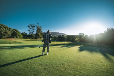 Téléchargez les photos : Une vue grand angle d'un homme afro-américain chauve éblouissant dans une élégante tenue taillée sur mesure ajustant ses lunettes de soleil tout en restant sur la pelouse d'un terrain de golf avec un club à la main sur un coucher de soleil - en image libre de droit