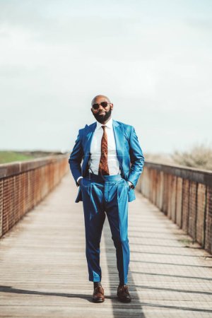 Photo for Selective focus on a tall bald African gentleman in a blue suit with a patterned brown tie to match his shoes; an Elegant black-bearded man wearing sunglasses and posing with his hands in his pockets - Royalty Free Image