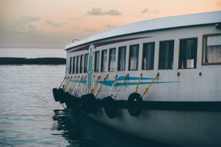 Foto de Un ferry de tamaño pequeño de color blanco con detalles azules amarrado al embarcadero y disparado al atardecer. Varios neumáticos de coche están unidos a ella por clips de metal que dejan rastros de óxido - Imagen libre de derechos