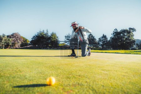 Téléchargez les photos : Un homme noir dandy portant des bretelles sur le dessus d'une chemise blanche, pantalon gris, et un chapeau avec des reflets bleus et rouges, s'agenouillant sur un terrain de golf vert luxuriant appuyé sur son putter et regardant la balle - en image libre de droit