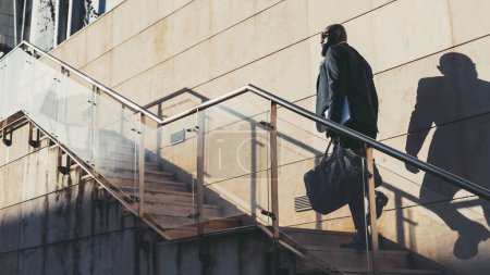 Foto de Un negro calvo con barba sube las escaleras para trabajar. El exitoso macho africano está subiendo una escalera en un día soleado, llevando un cuaderno y una bolsa de cuero con un traje gris y gafas de sol. - Imagen libre de derechos