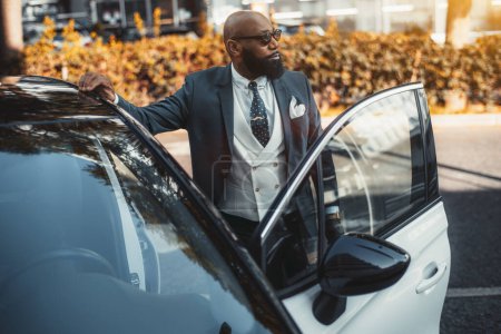 Photo for View of a handsome bald black man entrepreneur with a fine black beard, in sunglasses and a tailored fashionable suit, looking aside while entering his contemporary white car parked outdoors - Royalty Free Image
