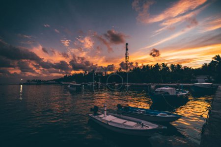 Téléchargez les photos : Vue en grand angle d'une jetée sur une île touristique tropicale avec un coucher de soleil atmosphérique en arrière-plan ; une jetée océanique avec quelques hors-bord et des ferries avec un paysage nuageux spectaculaire en soirée - en image libre de droit