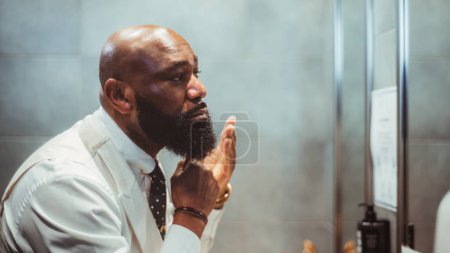 Photo for A profile view of a handsome bald black man grooming, brushing, and moisturizing the beard hair in front of the mirror in a bathroom of a luxury hotel; an African-American man taking care of his beard - Royalty Free Image
