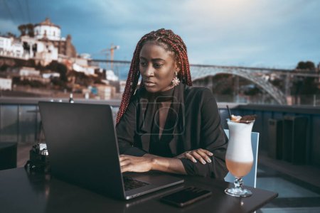 Foto de Un retrato de una mujer de negocios negra deslumbrante ocupada usando su computadora portátil mientras está sentada en un café callejero con un cóctel; una mujer empresaria afroamericana deslumbrante al aire libre con un netbook - Imagen libre de derechos