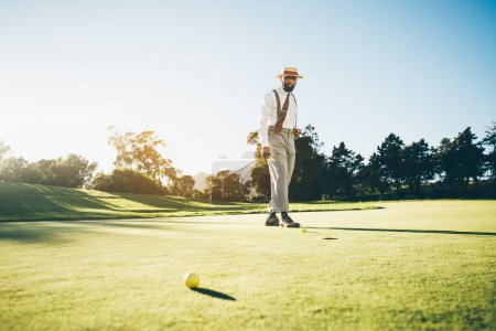 Photo for View of an elegant mature African American male golfer in a fashionable outfit with suspenders, holding a putter in his hand while walking across the golf course lit by the evening sun and thinking - Royalty Free Image
