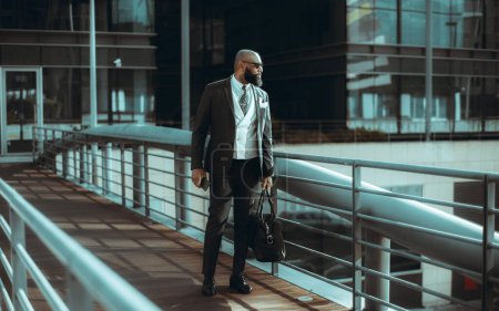 Photo for White-collar worker carrying a gym bag in one hand and a cell phone in the other. A bearded, bald black man wearing sunglasses, a dark suit, a tie and a white vest, exiting a contemporary building - Royalty Free Image