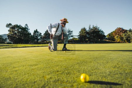 Photo for Bearded afro-descending man, all dressed up, wearing tailored grey pants, a white shirt, striped suspenders, with a hat. Kneeling on the floor of the golf course, leaning on his club, on a clear day - Royalty Free Image