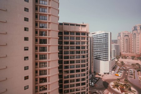 Téléchargez les photos : Grande hauteur de la ville à partir d'une chambre d'hôtel à Dubaï ; ville cosmopolite, où le ciel est bleu avec un paysage brumeux, gratte-ciel tout autour, et près d'eux un cercle de circulation avec des palmiers au milieu - en image libre de droit