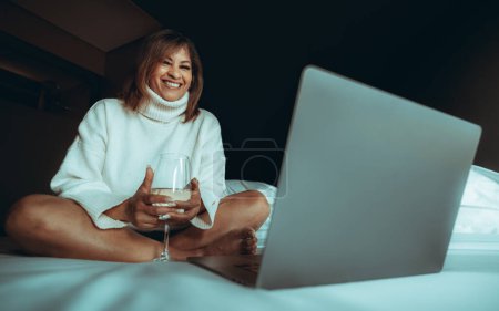 Foto de Una mujer hispana madura y hermosa con un jersey blanco de cuello alto está sentada en la cama en su acogedor dormitorio del hotel con una copa de vino blanco y navegando por la red en la computadora portátil. - Imagen libre de derechos
