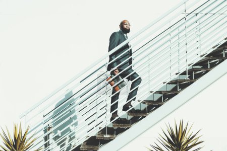 Photo for A bald black bearded man walks up the stairs. The African male is ascending a white metallic staircase on a shiny day, wears a dark suit and holds a leather purse in a very clean and cactus aesthetic - Royalty Free Image