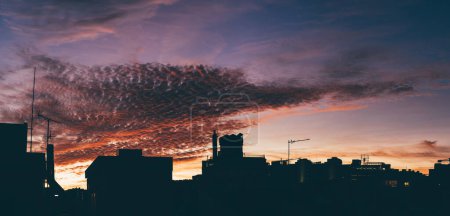 Foto de Un atardecer dramático crea un cielo atmosférico en tonos de azul, violeta y naranja, y en primer plano, una secuencia horizontal de siluetas de edificios residenciales con sus antenas de TV - Imagen libre de derechos
