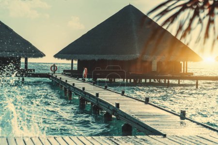 Téléchargez les photos : Une légende de confortables bungalows en bois délicats et aussi une passerelle en bois entourée d'eau cristalline dans une île paradisiaque ensoleillée aux Maldives, en arrière-plan un ciel nuageux et coloré pastel - en image libre de droit