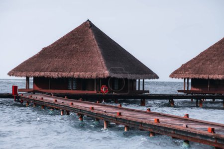 Photo for A walkway and a cottage made of wooden poles. The house has a pyramid hip roof, is over the crystalline turquoise waters of the Indian Ocean that lap gently at the deck of the luxury habitation - Royalty Free Image