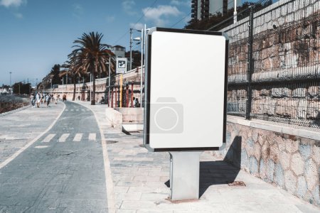 Téléchargez les photos : Un gabarit rectangulaire vertical de panneau d'affichage publicitaire blanc vide sur le trottoir ; une maquette d'un espace réservé à l'affiche vierge dans la promenade piétonne ; une maquette d'une bannière publicitaire urbaine près de la route - en image libre de droit