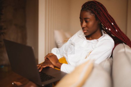 Photo for A portrait of a beautiful youth black female student with pink color box braids studying on her laptop while sitting on the beige couch in her living room, with a defocused background - Royalty Free Image