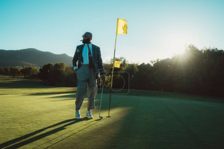 Ein bärtiger Mann mit Glatze und altem Geldstil trägt eine Sonnenbrille, raucht eine Zigarre und posiert an einem sonnigen Tag mit einem Golfschläger in der Hand neben einem Fahnenstock auf einem sattgrünen Golfplatz mit Sträuchern
