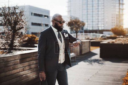 Téléchargez les photos : Un homme exécutif africain élégant debout dans un parc extérieur, vêtu d'un costume noir, d'un gilet blanc et de lunettes de soleil sombres vérifie l'heure sur sa montre-bracelet avec des bâtiments modernes à l'arrière - en image libre de droit
