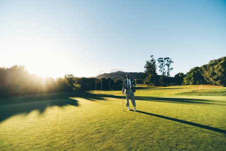 Foto de Una captura de un hombre africano, con un estilo hipster, posando con un palo de golf en su mano en un día soleado, cielo despejado, en un campo de golf verde exuberante con arbustos y la bengala del sol en su espalda - Imagen libre de derechos