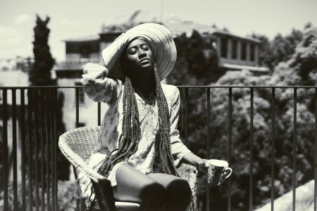 Foto de Foto en blanco y negro de una joven de origen afrodescendiente, vestida de manera femenina, con un vestido florido, un sombrero de ala ancha y pelo largo trenzado, en la azotea de un apartamento mientras toma el sol. - Imagen libre de derechos
