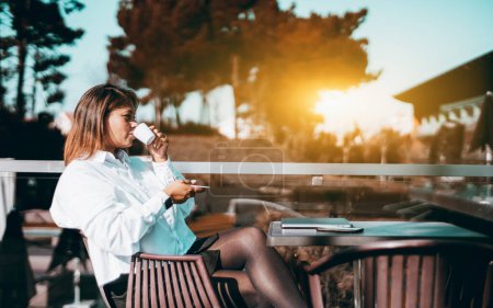 Photo for Profile photo of a Brazilian woman having a coffee in an establishment with an outdoor terrace made of wood surrounded by trees and with sunbeam and clear sky in the background - Royalty Free Image