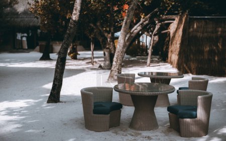 Foto de Set de muebles de jardín, para comidas, en plástico marrón oscuro imitando madera. Se asientan en la playa de arena blanca, con árboles circundantes y edificios de hojas de palma - Imagen libre de derechos